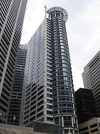 Ground-level view of a narrow, 30-storey skyscraper with a rectangular cross section and rounded anterior siding; a protruding circular spire is visible on the roof.