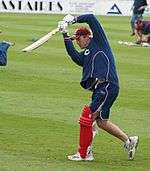 A man wearing dark blue shorts and sweat-shirt with a large red pad fastened to his left leg holds a cricket bat in front of him.
