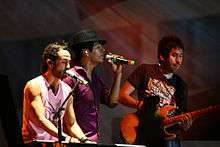 Three men singing. The left has a sleeveless shirt, is playing a keyboard, and sings in a microphone stand. The men at center is wearing a purple shirt and has a black hat and is holding a microphone. On the right, there is another man, wearing a black shirt with a white legend, holding an electric guitar.
