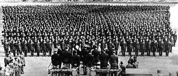 In the foreground a platform with officers facing away from the camera. In the background a formation of over a thousand soldiers, raising their right arms