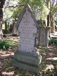 An elegant sandstone gravestone with a gabled top, standing in dappled light under trees
