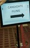 A sign with a black frame holds a piece of paper reading "CANDIDATE FILING" with an arrow pointing to the right