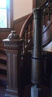 An old brass cannon mounted vertically next to a curved stairway in a lobby with wooden paneling.