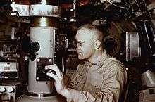 Captain Edward L. Beach dressed in khaki uniform gazing through the viewfinder while adjusting the focus using dial control on the periscope in the conning tower of the nuclear submarine USS Triton