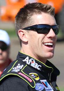 Head and shoulders of a man in his early thirties. He is facing to the right, smiling and wearing black sunglasses. He is wearing black racing overalls, with the Aflac logo embroiled on his collar with logos for Ford, Valvoline, Simpson and NASCAR displayed on the body.