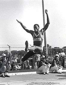 A man midway through a long jump leap.  There is a metal chainlink fence in the background, both in front of which and behind are a number of spectators