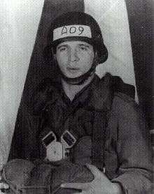 A black-and-white photo depicting Lozada from the waist up holding a sleepeing back in his hands, wearing a parachute and a Kevlar helmet with the letters A09 on it.