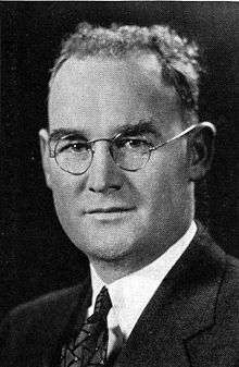 Quarter-length black-and-white image of a man. He is wearing a jacket and tie, and his hair is short. He is wearing spectacles