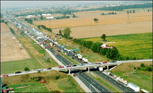 "A highway viewed from high above travels into the distance from the bottom-right to the top-left. An overpass allows a road to cross the highway near the bottom of the image. The surroundings are entirely agricultural. On the highway, several dozen vehicles are piled into each other. The middle of the large pileup is smoking."