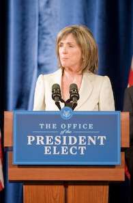 A pale-skinned woman in her fifties with straight, light brown hair parted near the middle and falling to above the shoulders, wearing a light beige suit jacket, listening with a serious expression to an unseen question as she stands behind a brown podium with a blue and white sign saying "Office of the President Elect" and two black microphones, against a blue drapes background.