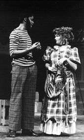 black man with sideburns, striped shirt, and plaid pants talks to white girl in calico dress with hat and purse on a stage