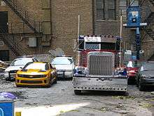 A Peterbilt 379, and a yellow Camaro are beside each other, with two police cars behind. They are near a building complex.