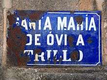 A very rusty metal sign covered in blue enamel with white enamel lettering, the sign affixed to a stone wall. The words on the sign are "Santa María de Óvila Trillo".