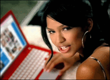 Image shows a bed with a switched-on red laptop. Beside the laptop lies a dark-haired woman in a sleeveless top, wearing large circular earrings while her hair is tied up with a fringe covering the left side of her face.