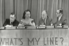 Three men and one woman sit at a long table. Behind them is a curtain. On the from of the table are the words "What's My Line?".