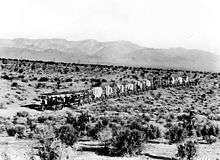 A train of over fifteen wagons crossing the desert, a flat sandy expanse with many scattered scrub-like bushes and a mountain range in the distance.