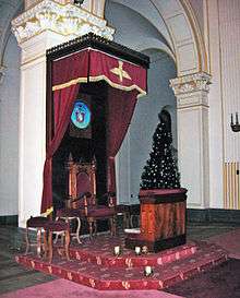 A polished wooden chair on a raised dais, under a polished wooden canopy trimmed with crimson curtains