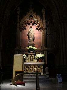 A starkly lit stone alcove with a pointed arch and a statue of a man in a robe with one hand over his chest. Below are shelves with bright golden objects on them. A small folded partition and low chair is on the floor at left