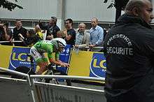 A road racing cyclist, wearing a green skinsuit and an aerodynamic helmet sitting low and crouched on his bicycle. Spectators watch from behind roadside barricades.