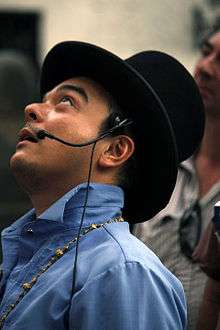 A head shot of Carlos Celdran wearing a top hat, headset microphone, and blue barong, looking skywards as he speaks.