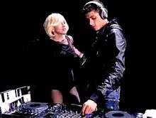 A blond woman and a brunette man facing each other against a stark black background. The man wears a navy blue jacket and has headphones on his ears. With his left hand he is busy operating a DJ's music console. The woman has short blond bob cut hair and wears a shot black dress. She is looking towards the man.
