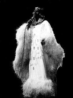 A woman holding a microphone, looking to the side, wearing a white dress with feathers.