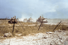 Four main battle tanks on a road kick up dust as they move towards a defensive position.