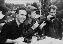 Three men are sitting at a small table. Man at left is holding sunglasses in his right hand, smiling, leaning forward and looking to his right. Man in middle has elbows on a brief case, gesturing with upraised hands, right hand is holding sunglasses, he is looking to his left. Third man has a small cup held to his lips by his right hand.