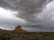 Dark, rolling storm clouds lower over a desert landscape; a butte stands in the near distance, left of center.