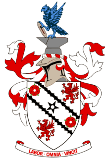A white shield upon which emblems of two red roses and two red griffins appear at alternate corners. A black five-pointed star is in the centre of the shield's design between two black diagonal lines. Around the shield are red and white ribbons in a symmetrical design. Above the shield is a silver-coloured knight's helmet surmounted by grey anvil upon which a vivid azure-coloured eagle is perched, holding a grey shuttle. Below the shield is the motto "LABOR OMNIA VINCIT".