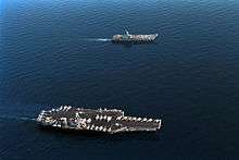 Aerial photograph of two aircraft carriers sailing in concert on calm water. The upper ship is smaller, and has a small number of aircraft on its flight deck. The larger carrier, with a flat deck crowded with planes and helicopters, is towards the bottom.