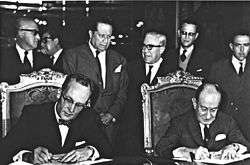 A black and white photograph of two mature and formally dressed men seated in chairs, signing some papers over a large table while a group of six men in suits stand behind their seats.