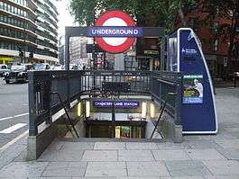 A staircase leading down below a sidewalk with a sign above reading "UNDERGROUND" in white letters on a blue rectangle in front of a red circle