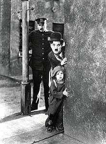 A black and white image of a young man with a mustache and a bowler hat peaking around from behind a brick corner with a police officer behind them