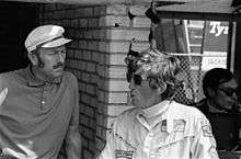 Black-and-white photograph of Colin Chapman on the left and Rindt on the right conversing in the pit lane in front of a brick wall