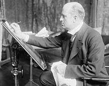A man in late middle-age, facing left, drawing at an upright drawing board.