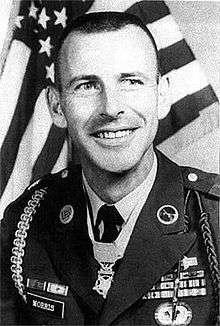 A black and white image of Morris from the chest up in his military dress uniform with ribbons. He has no hat and there is an American flag in the background.