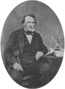 A middle aged Victorian gentleman sits beside a table wearing a dark suit with waistcoat and bow tie. His left elbow rests on the table. In his right hand he holds a quill pen.