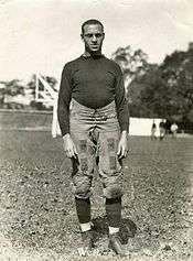 An African American male in an 1920s football uniform stands on a football field. Several unidentifiable individuals are in the visible in the distance