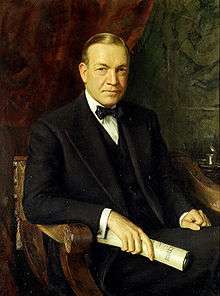 Man of about 65 in an ornate wooden chair. Formally dressed in a dark suit, white shirt, and black bow tie, he holds a rolled document in his left hand.