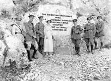 Five soldiers in Sam Browne belts, riding boots and peaked caps, and one woman in light coloured dress and matching hat in front of a stone inscription that reads: "The British XXI Corps with Le Detachment Français de Palestine et Syrie occupied Beirut and Tripoli October 1918 AD."