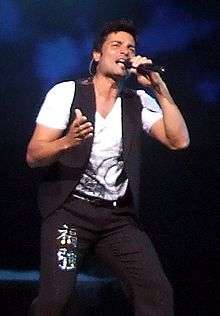 A man with a white T-shirt and a black vest singing into a microphone.