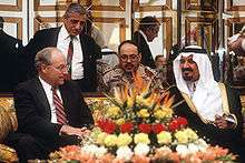 A balding man with glasses, wearing a suit, sits on the left of a couch next to a hirsute man in traditional Arab headress and attire on the right. In front, on a coffee table, is a vase full of flowers; behind them is a man in desert camouflage and another man in a suit in front of a mirrored partition