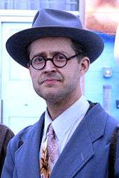 Photograph of a man in a formal suit, hat, and glasses