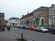 Street scene showing road junction and grey stone buildings with parked cars in front of them. To the left is a grassy area with a tree.