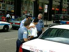 A man on a city street in a white shirt with his hands cuffed behind his back, with three police officers behind him. The one at rear is opening the door of the police car in the bottom right of the image.