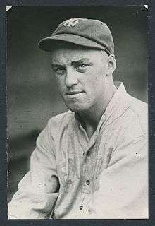 A man wearing a white baseball jersey and dark cap with a white "NY" on the center looks into the camera.