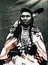 American Indian male sitting with one feather in hair and wrapped in a patterned blanket