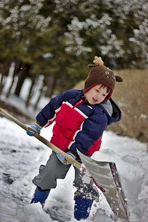 A child in appropriate clothing, moving a shovelful of fresh snow