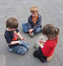 Three children aged about six years are in a group on the ground, a boy and girl kneeling and another boy seated cross-legged. The two kneeling children hold marbles. There are other marbles in a bag on the ground. They appear to be negotiating over the marbles. The third child is watching.
